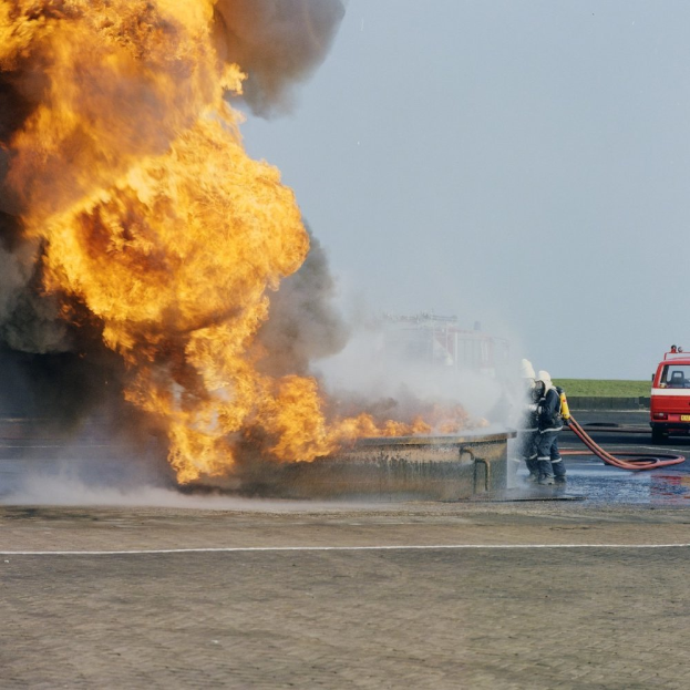 Ein Feuerwehrauto steht in Flammen am Rande einer Straße, mit zwei Personen in Helmen und Schlöschen in der Nähe und einem Fahrzeug und dem Himmel im Hintergrund.