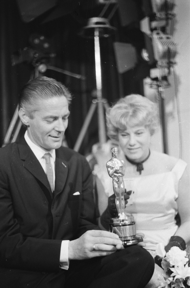 Ein Mann in einem schwarzen Blazer, wei├čem Hemd und Krawatte hölt eine Oscar-Statue, waehrend er neben einer Frau in Abendgarderobe sitzt, die einen Blumenstrau├č hölt, mit Hintergrundbeleuchtung in einem Schwarz-Wei├č-Bild.