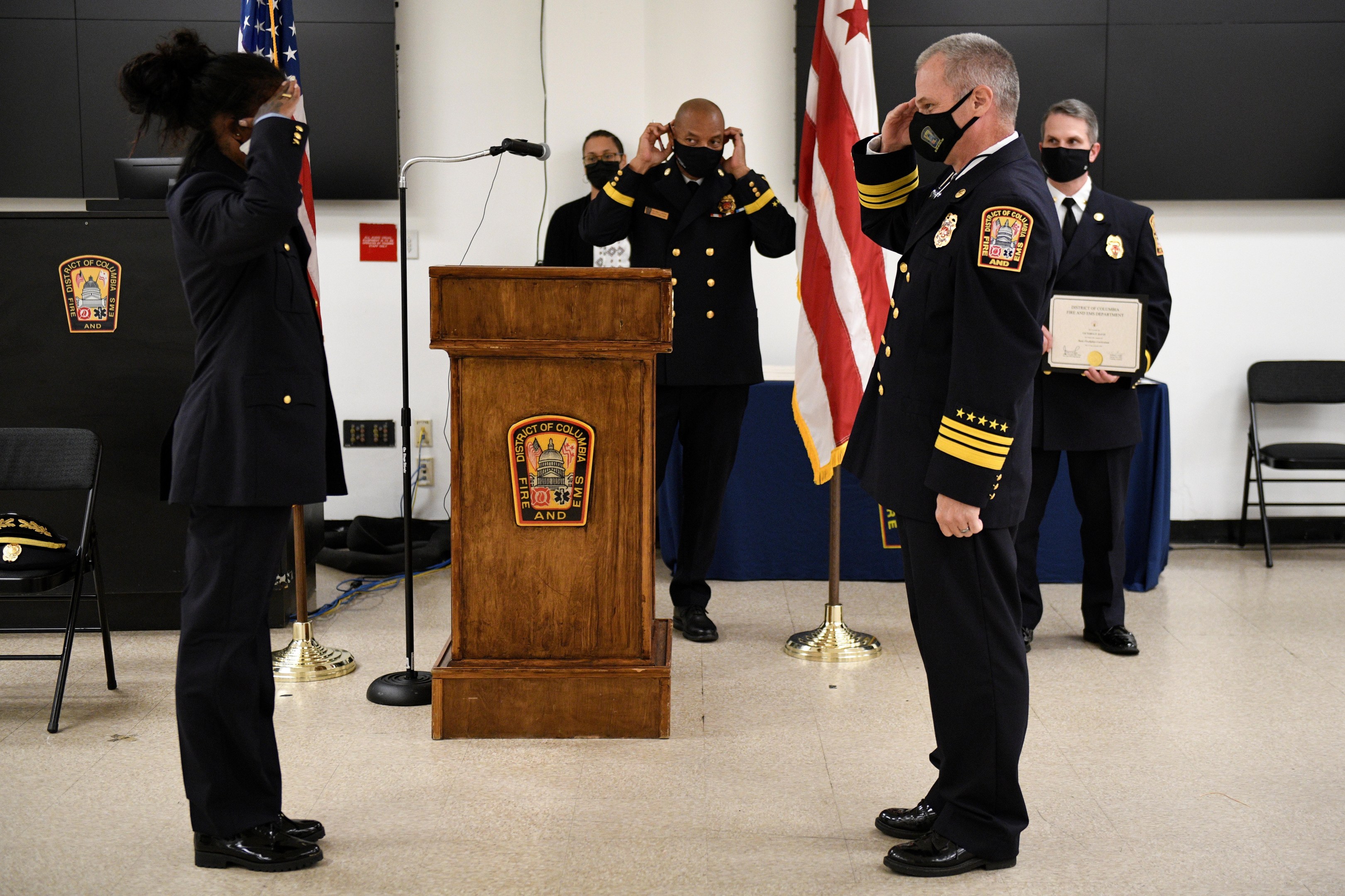 Gruppe uniformierter Personen auf einem Podium während einer Vereidigung eines neuen Feuerwehrchefs, der eine Urkunde hält, mit Flaggen, Stühlen, Tischen und einer Wand im Hintergrund.