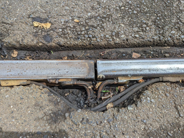 Nahaufnahme eines Metallkanalrohrs am Straßenrand mit angebrachten Drähten und trockenen Blättern in der Nähe.