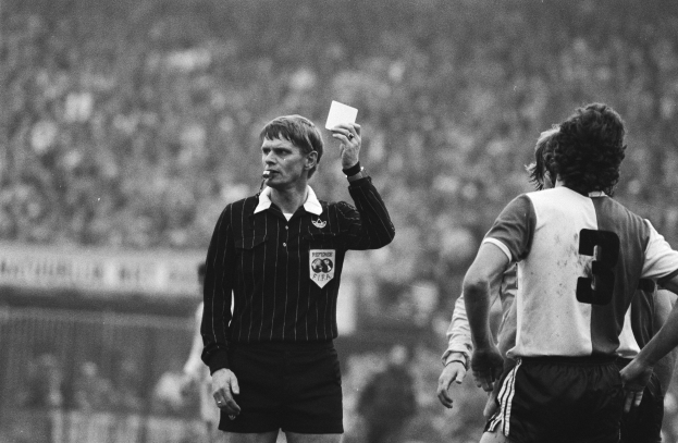 Schwarzes und weißes Bild eines Schiedsrichters, der eine rote Karte vor einer Menge in Fußballuniformen hält, mit einem Zaun im Hintergrund.