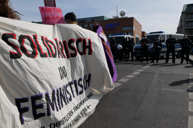 Eine Gruppe von Menschen marschiert auf einer Straße, wobei sie ein Banner tragen, auf dem 'Solidarität und Feminismus' steht, mit parkenden Fahrzeugen und Gebäuden im Hintergrund unter einem klaren blauen Himmel.