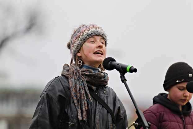 Eine Frau in schwarzer Jacke, Schal und Mütze singt in ein Mikrofon auf der Bühne, mit einer anderen Person in einer maronenfarbenen Jacke und schwarzer Mütze rechts von ihr, vor einem unscharfen Hintergrund.