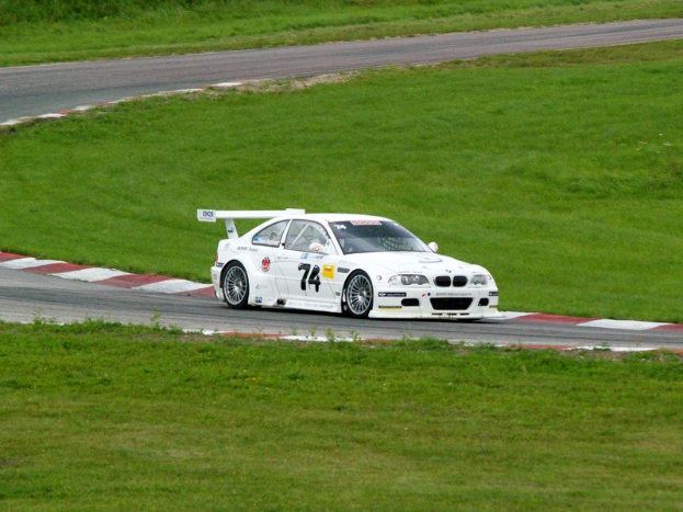 Ein weißer BMW M3 GT3 Rennwagen fährt auf einer Rennstrecke mit grünem Gras und Pflanzen im Hintergrund.