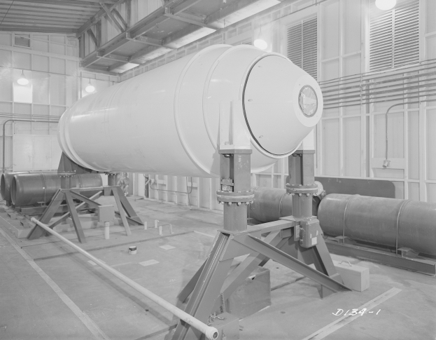 Ein Schwarz-Weiß-Foto einer großen zylindrischen Wasserstoffbombe auf einem Ständer in einer industriellen Fabrikumgebung mit sichtbaren Rohren, Beleuchtung und Text unten.