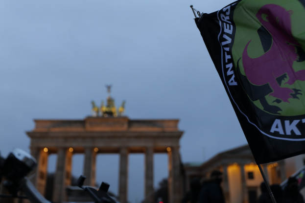 Eine Gruppe von Menschen steht vor dem Brandenburger Tor in Berlin, Deutschland, mit einem Fahnenmast, der ein Logo und Text im Vordergrund trägt.