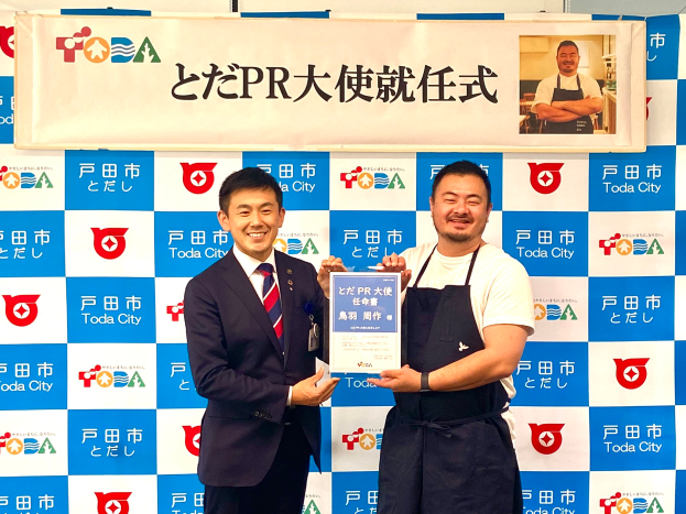 Zwei lachende Männer mit einem Zertifikat, mit einem Banner im Hintergrund, auf dem "Toda City Wins PR Award" steht und ein Foto einer Person zeigt.