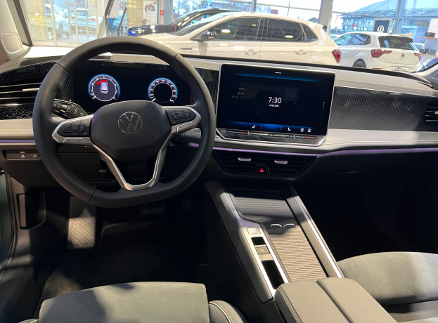 Das Innere eines Volkswagen e-Golf mit einem Lenkrad, Armaturenbrett, Bildschirmen, Knöpfen, Sitzen und Glastüren, mit Blick auf andere Autos, Gebäude, Bäume, Pfosten und den Himmel durch die Glastüren.