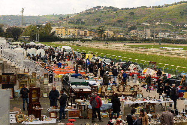 Große Gruppe von Menschen auf einem Flohmarkt mit Tischen, auf denen Gegenstände wie Foto Rahmen und Stühle ausgelegt sind, umgeben von geparkten Fahrzeugen, Geländern, Stufen, Bäumen, Gebäuden, Laternenmasten, Hügeln und einem bewölkten Himmel.
