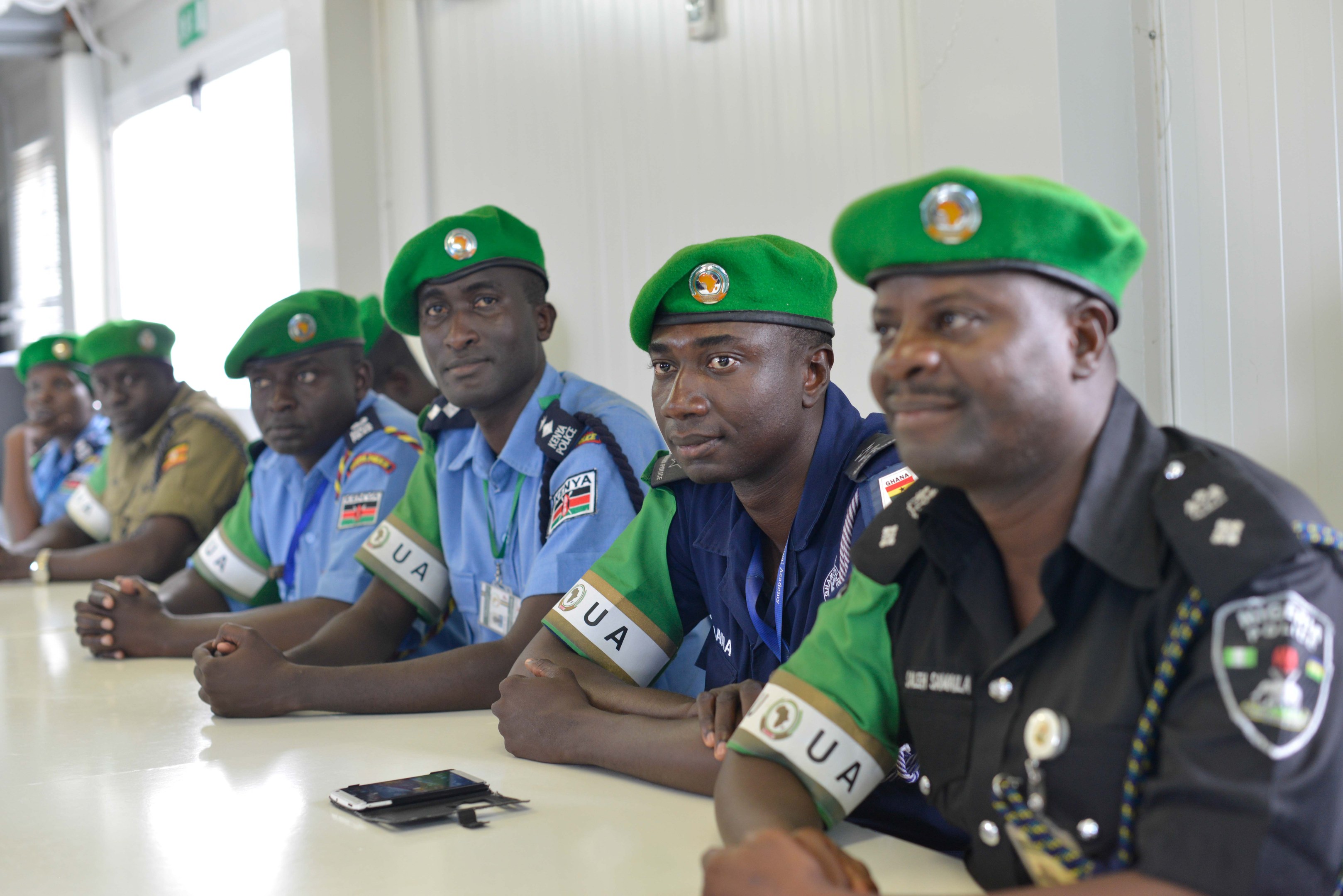 Gruppe von Polizeibeamten sitzt an einem Tisch während einer Schulung, mit Mobiltelefonen und anderen Gegenständen auf dem Tisch, in einem Raum mit Fenstern und Deckenbeleuchtung.