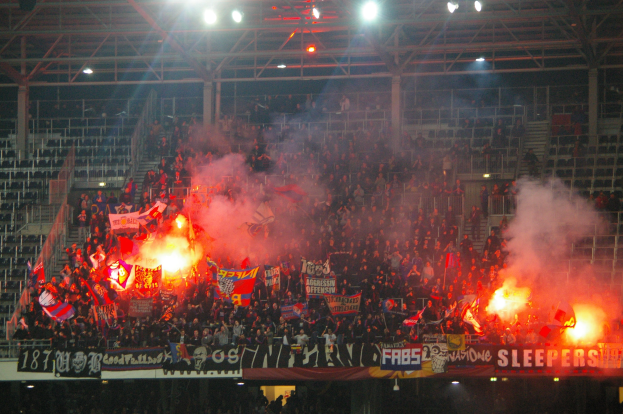 Eine große Menschenmenge in einem Stadion hält Fahnen und Banner, während Raketen und Rauch die Luft erfüllen und Banner, Metallrahmen, Stangen und Deckenleuchten zu sehen sind.