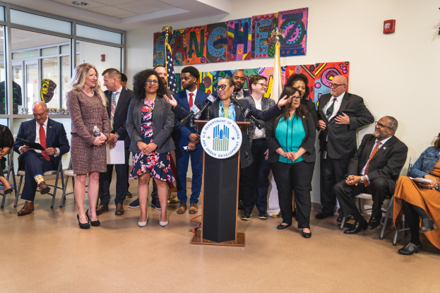 Gruppe von Menschen vor einem Rednerpult mit einem Mikrofon und einer Tafel mit der Aufschrift "Sancho Stadtrat tritt dem Stadtrat bei", mit sitzenden und stehenden Zuschauern, Flaggen, einem Feuerl├Âscher und Deckenleuchten im Hintergrund.
