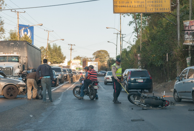 Eine Gruppe von Menschen steht neben einem verunglückten Motorrad am Straßenrand mit mehreren Fahrzeugen, Bäumen, Polen und Lampen im Hintergrund.
