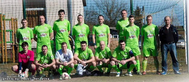 Eine Gruppe von Männern in grünen T-Shirts steht auf einem Fußballfeld mit einem Ball und einem Tor, umgeben von Bäumen, Gebäuden und Fahrzeugen.