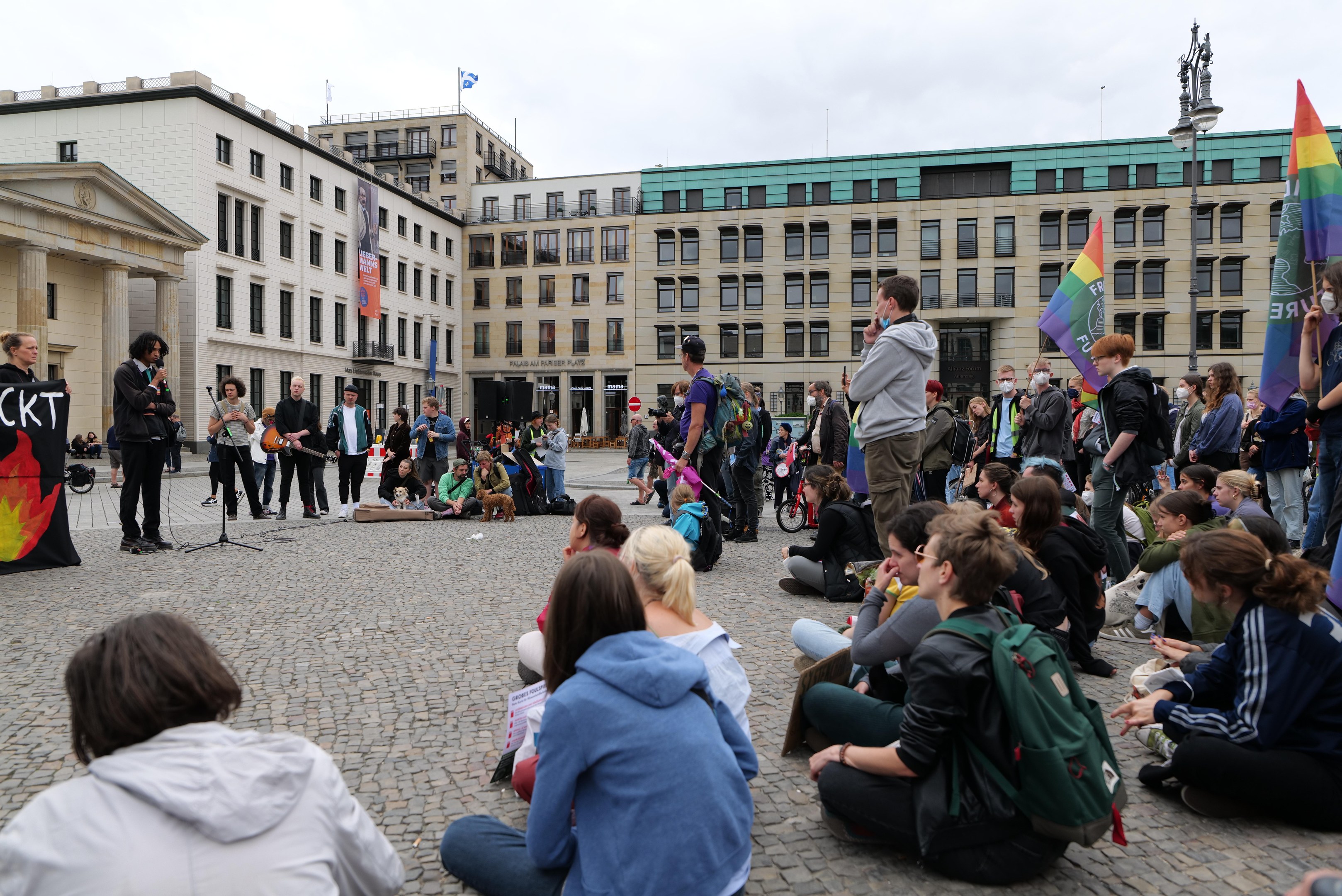 Eine Gruppe von Menschen, die auf dem Boden vor einer Menge sitzen, die Fahnen und Plakate hält, mit einer Person, die in ein Mikrofon spricht, einer Statue und Gebäuden im Hintergrund während einer anti-schwulen Demonstration in Berlin.