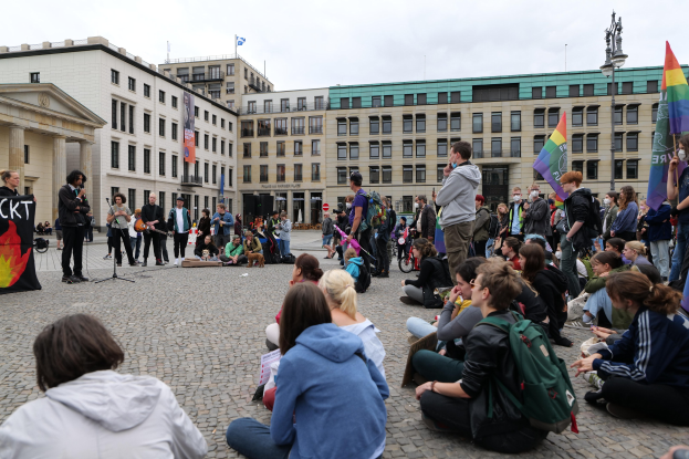 Eine Gruppe von Menschen, die auf dem Boden vor einer Menge sitzen, die Fahnen und Plakate hält, mit einer Person, die in ein Mikrofon spricht, einer Statue und Gebäuden im Hintergrund während einer anti-schwulen Demonstration in Berlin.