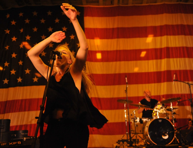 Frau singt in ein Mikrofon auf der Bühne mit einem amerikanischen Flaggenhintergrund, begleitet von einem Schlagzeuger, mit Bühnenausrüstung und Beleuchtung im Hintergrund.