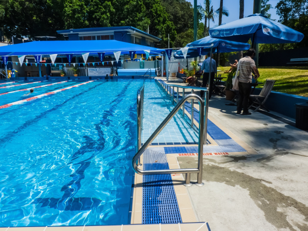 Ein großes Schwimmbad mit Schwimmern, umgeben von Geländern, Sonnenschirmen, Stühlen, Tischen, Grünpflanzen und einem Gebäude unter einem klaren blauen Himmel.