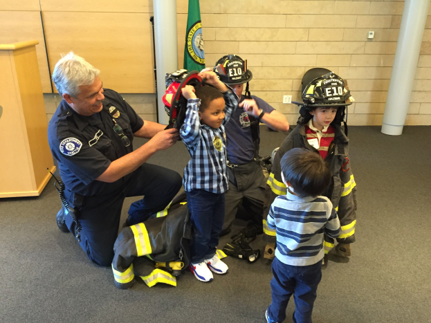 Eine Gruppe von Kindern in Feuerwehruniformen und ein Feuerwehrmann in Uniform stehen um ihn herum, während er einen Helm hält, mit einer Tasche auf dem Boden in einer Feuerwache.