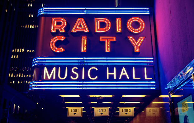 Das erleuchtete Radio City Music Hall in New York City mit hohen Gebäuden im Hintergrund, das eine 'Radio City' -Leuchtschrift und zusätzliche Schilder auf der rechten Seite zeigt.
