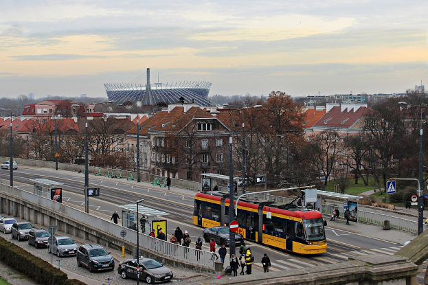 Eine gelbe und schwarze Straßenbahn fährt auf einer Stadtstraße mit hohen Gebäuden, umgeben von Autos, Fußgängern, Laternen, Schildern, Bäumen und Gras, mit bewölktem Himmel im Hintergrund.