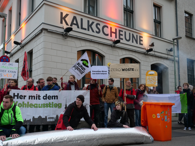 Eine Gruppe von Menschen mit Protestschildern vor einem Gebäude, daneben weitere Sitzende und ein Mülltonne auf der rechten Seite, in einer deutschen städtischen Umgebung.