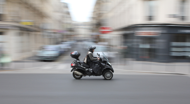 Eine Person in schwarzer Kleidung und Helm fährt mit anderen Fahrzeugen, Gebäuden und einem klaren blauen Himmel im Hintergrund auf einer Stadtstraße mit einem Motorroller.