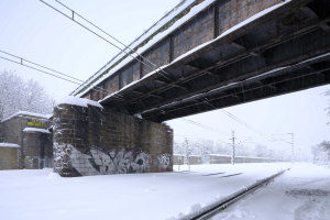 Eine schneebedeckte Zugstrecke unter einer Brücke mit Graffiti, Strommästen, Bäumen und einem Himmel im Hintergrund.