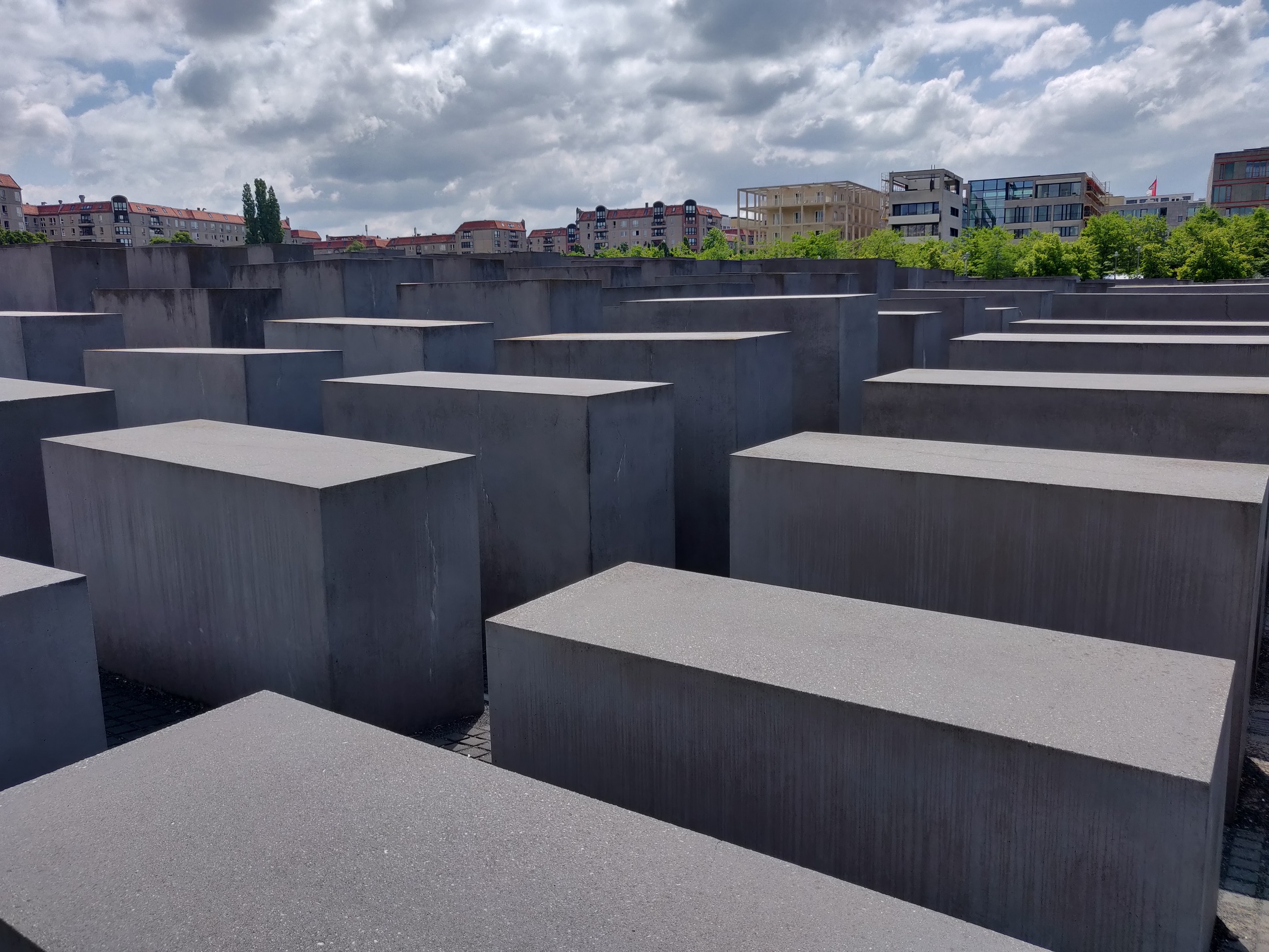 Konkreter Holocaust-Mahnmal mit rechteckigen Blöcken in einem Gittermuster umgeben von Bäumen, Gebäuden und einem bewölkten Himmel.