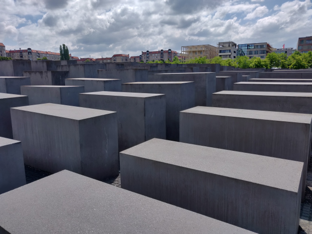 Konkreter Holocaust-Mahnmal mit rechteckigen Blöcken in einem Gittermuster umgeben von Bäumen, Gebäuden und einem bewölkten Himmel.