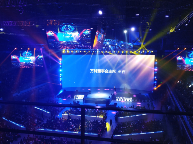Menschenmenge vor einer hell erleuchteten Bühne mit Metallabsperrungen, die auf ein E-Sport-Event warten, während auf der Bühne Performer stehen und ein großer Bildschirm im Hintergrund angezeigt wird.