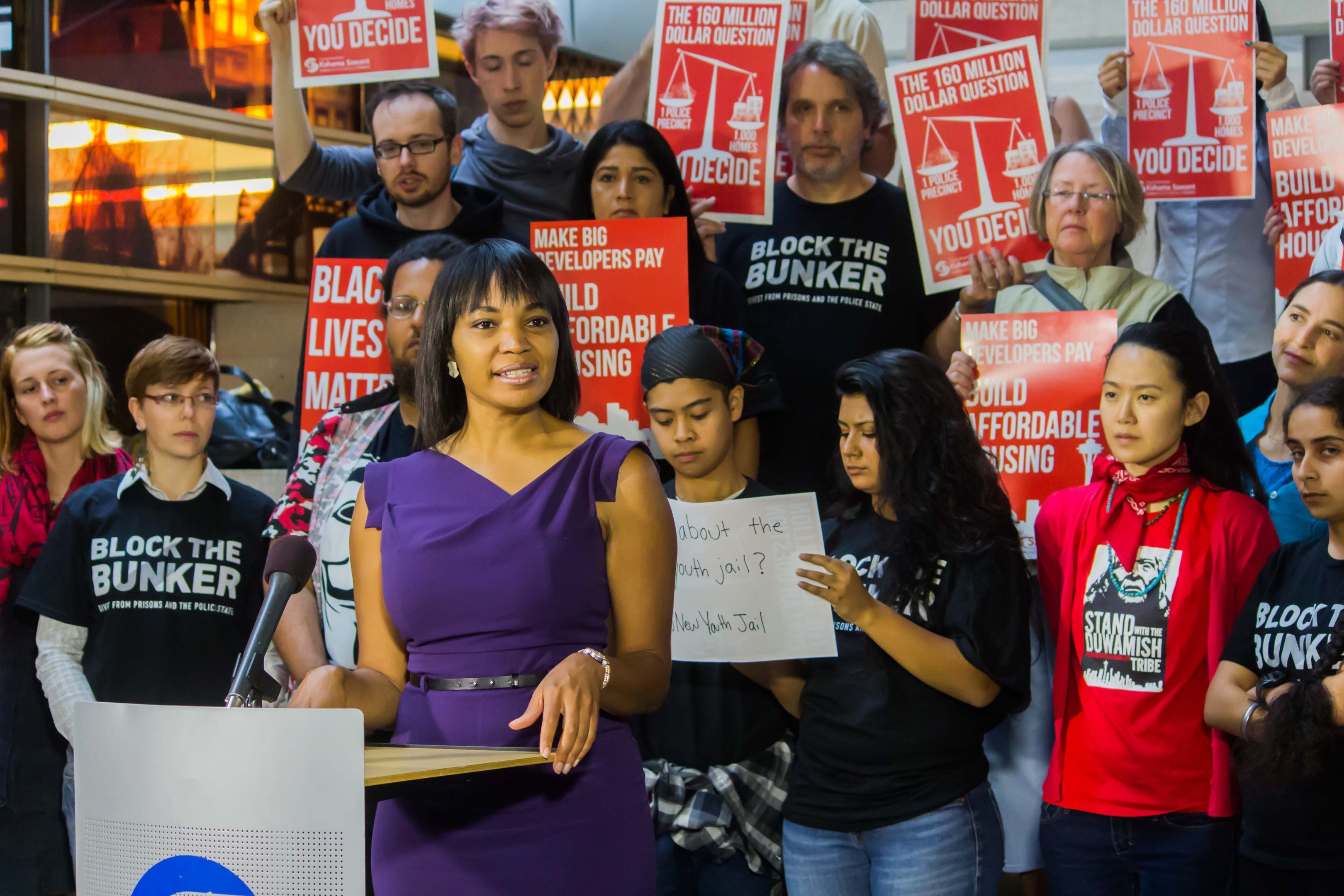 Black Lives Matter-Aktivistin spricht vor Publikum mit Mikrofon, das Publikum hält Schilder, im Hintergrund gibt es Lichter und Gegenstände.