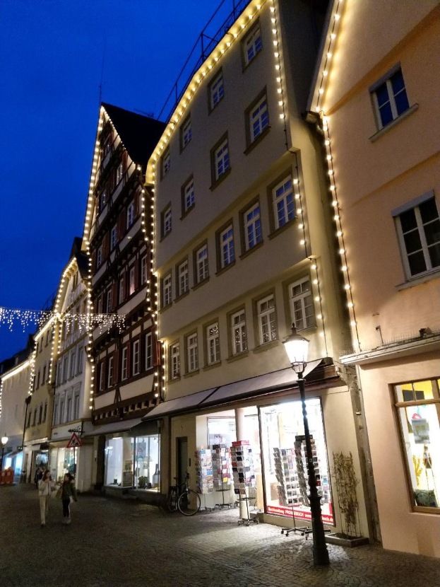 Headsteinstraße in Rothenburg ob der Tauber, Deutschland, beleuchtet von festlichen Weihnachtslichtern mit Menschen und einem geparkten Fahrrad, flankiert von Fenstern und einem sichtbaren Himmel.