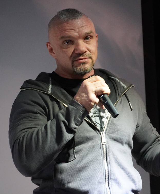 Ein Mann in einer schwarzen Kapuzenjacke und einem schwarzen Anzug mit weißem Hemd, der ein Mikrofon hält und selbstbewusst vor einer Wand steht.