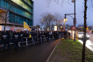 Große Gruppe von Menschen mit Schildern vor einem Gebäude, mit Barrikaden und Bäumen während einer Demonstration in Berlin.