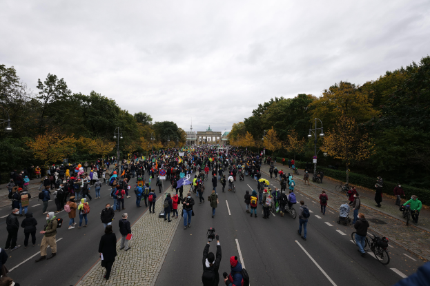 Eine große Gruppe von Menschen, die eine von Bäumen gesäumte Straße in Berlin entlanggehen, einige halten Kameras, mit einem Gebäude im Hintergrund und einem klaren Himmel darüber.