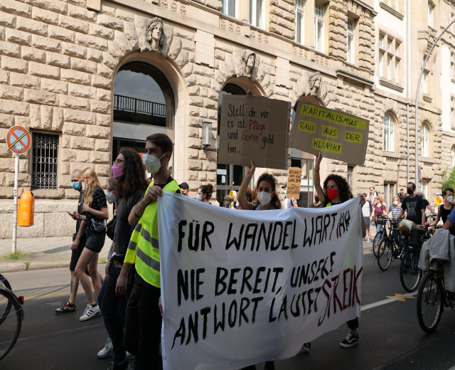 Eine Gruppe von Menschen, die während einer Demonstration in Berlin die Straße entlanggehen, einige mit Plakaten und Transparenten, andere mit Fahrrädern, mit einem Gebäude im Hintergrund, das Fenster, Bögen, Säulen und Skulpturen aufweist.