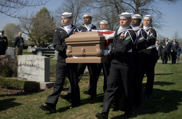 Gruppe uniformierter Männer in weißen Mützen und schwarzen Uniformen trägt einen Sarg über eine grüne Friedhofswiese mit Bäumen, umgeben von Menschen, Autos und einem klaren blauen Himmel.