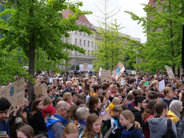 Eine große Menschenmenge protestiert vor einem Gebäude in Berlin, mit Schildern, Bäumen, Fahrzeugen, einem Lautsprecher und Himmel.