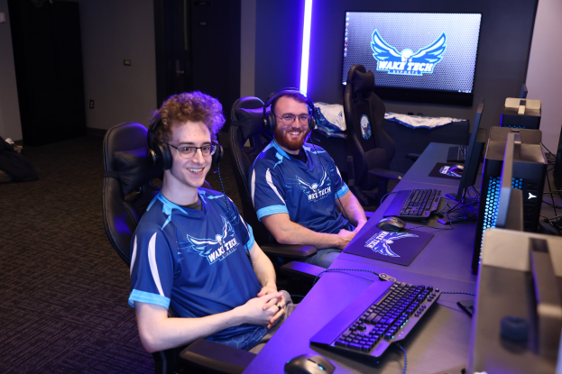 Zwei Männer mit Headsets sitzen an einem Schreibtisch mit Monitoren, Tastaturen und anderem Equipment und lächeln beim Spielen eines Spiels; ein Bildschirm und Lichter sind im Hintergrund sichtbar.