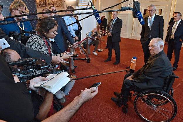 Ein Mann im Rollstuhl spricht mit Reportern, die Kameras, Mikrofone und Handys halten, mit einem Banner und Lichtern im Hintergrund.