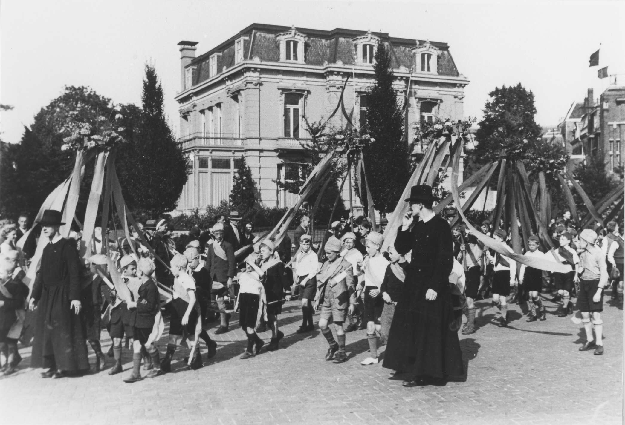 Ein Schwarz-Weiß-Foto einer Gruppe von Menschen, die eine Straße entlanggehen, einige halten Fahnen, mit Hüten, Bäumen, Gebäuden und einem sichtbaren Himmel im Hintergrund, mit einem Maypole als Indikator für ein Maifeier.