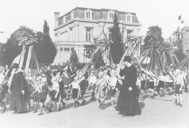 Ein Schwarz-Weiß-Foto einer Gruppe von Menschen, die eine Straße entlanggehen, einige halten Fahnen, mit Hüten, Bäumen, Gebäuden und einem sichtbaren Himmel im Hintergrund, mit einem Maypole als Indikator für ein Maifeier.