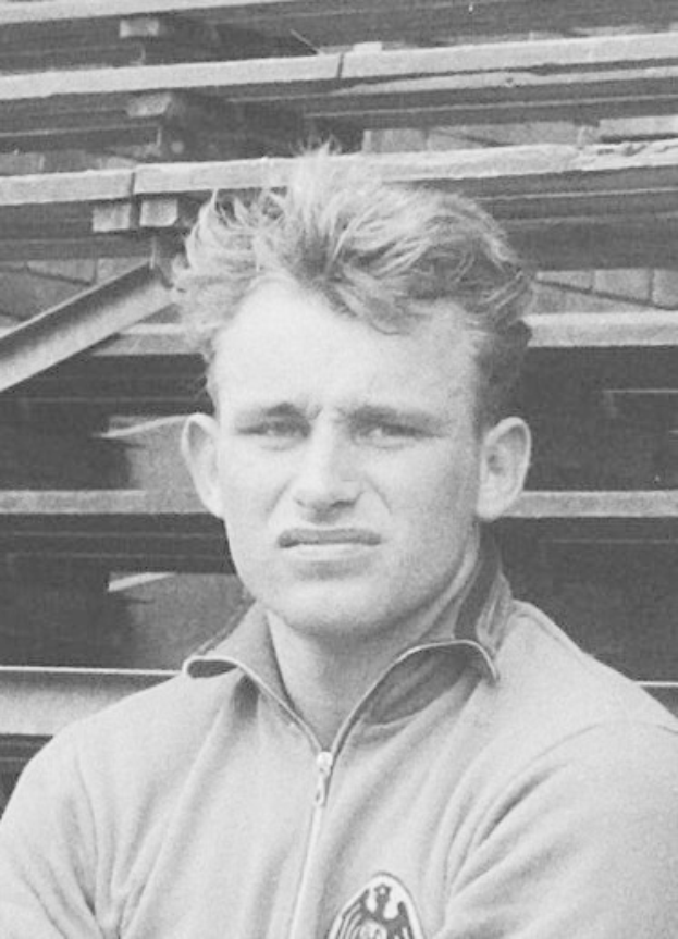 Schwarzes und weißes Foto eines Mannes in einer Jacke vor einem Stapel Holzplanken, der als legendärer deutscher Fußballspieler gilt.