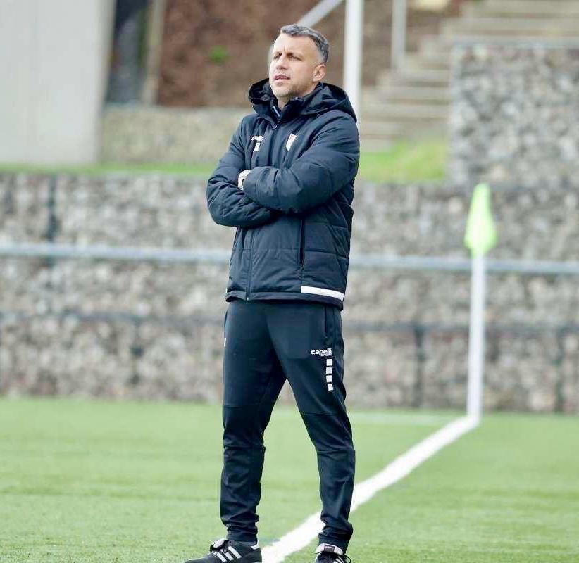 Ein Mann in schwarzer Jacke und Hose steht auf einem Fußballfeld mit verschränkten Armen und sieht aus wie ein Trainer während eines Trainings.