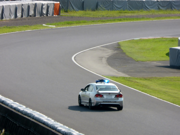 Polizeiauto auf einer Rennstrecke mit Gras auf beiden Seiten, Menschen, Zelte und andere Gegenstände im Hintergrund.