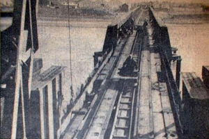 Ein Schwarz-Weiß-Foto einer Bahnschiene mit einer im Hintergrund befindlichen Brücke, umgeben von Werkzeugen und Materialien.