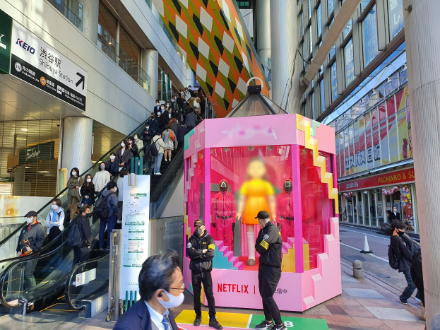 Gruppe von Menschen vor einem Gebäude mit Glaswänden mit Texttafeln, einige tragen Masken, in der Nähe einer Rolltreppe; ein pinker Kasten in der Mitte zeigt "Netflix's new Netflix-Booth in Tokyo" mit Schaufensterpuppen drin.