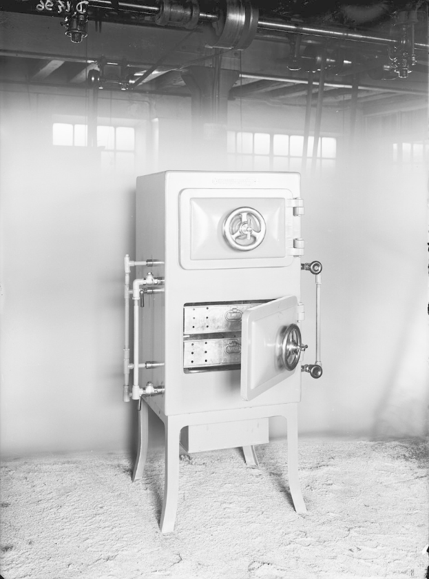 Schwarzes und weißes Foto eines altmodischen Safes in einem Raum mit einer Wand, Fenstern und an der Decke verlaufenden Rohren.