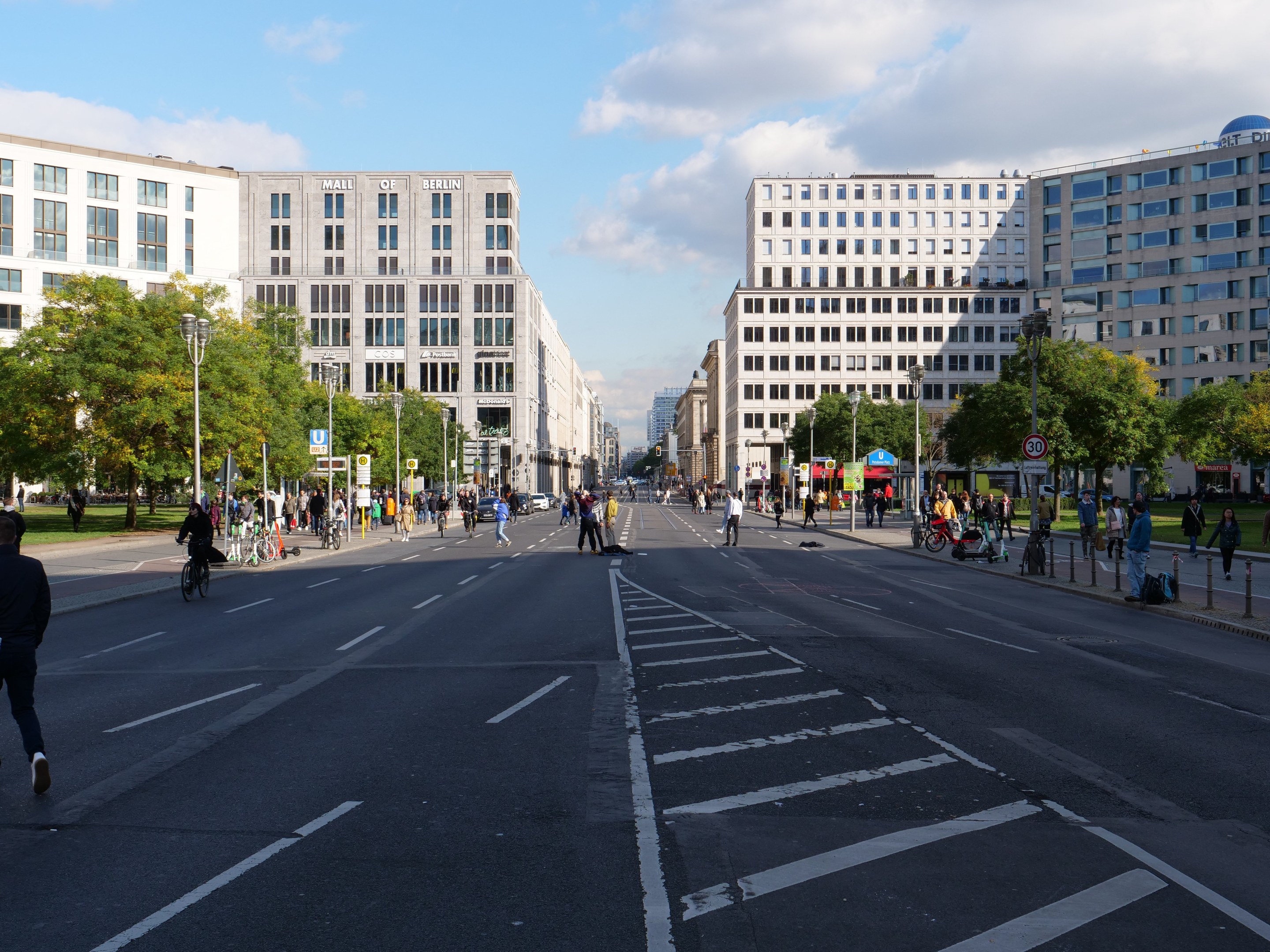 Eine belebte Stadtstraße in Berlin, Deutschland, mit Fußgängern und Radfahrern auf der Straße, hohen Gebäuden, Bäumen, Straßenlaternen und Schildern unter einem bewölkten Himmel.